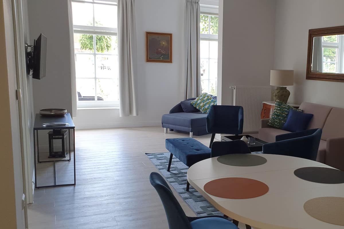 A spacious living room with a large window, a sofa, a dining table with chairs, and a TV mounted on the wall.