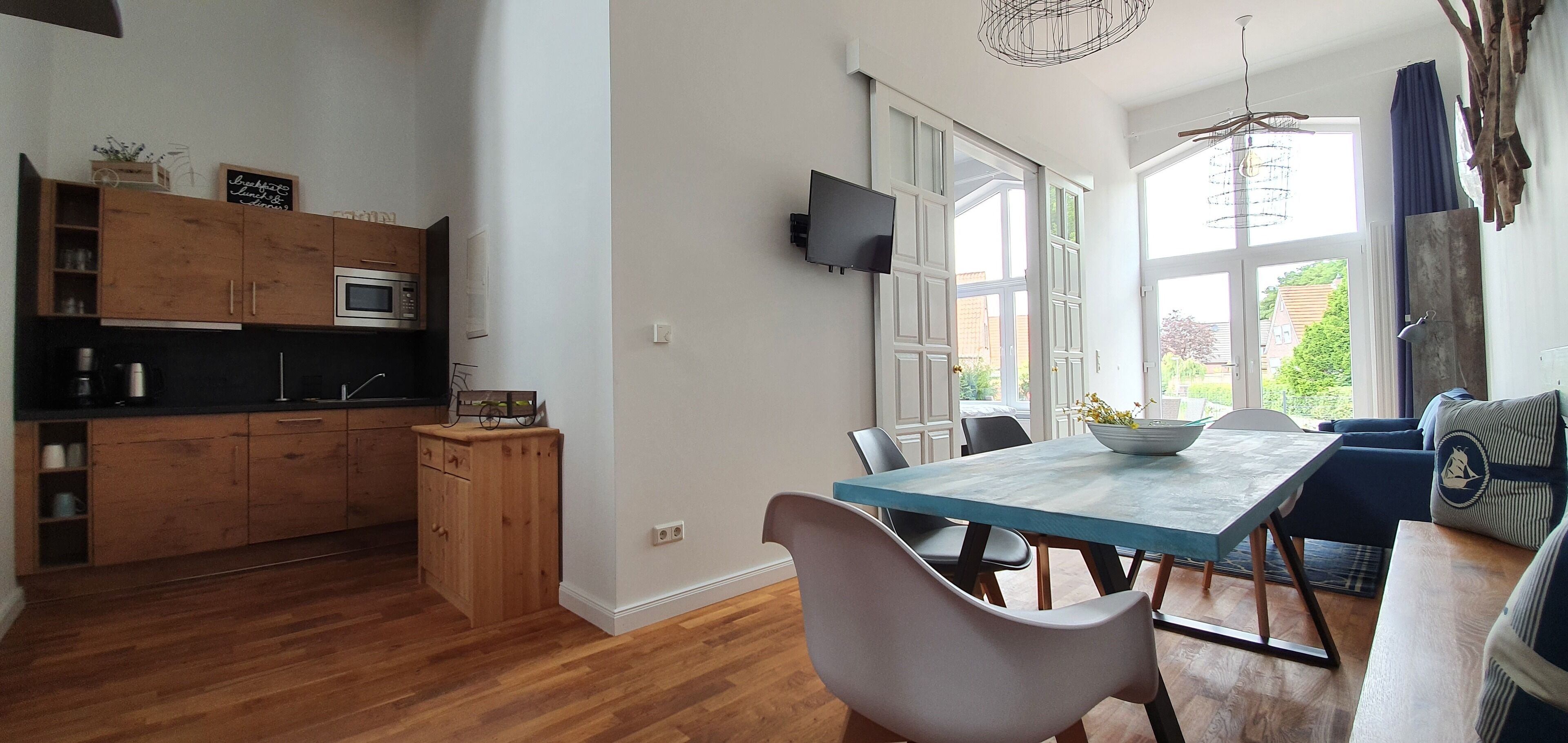 A modern kitchen with wooden cabinets, a dining area with a blue table, and a TV mounted on the wall.