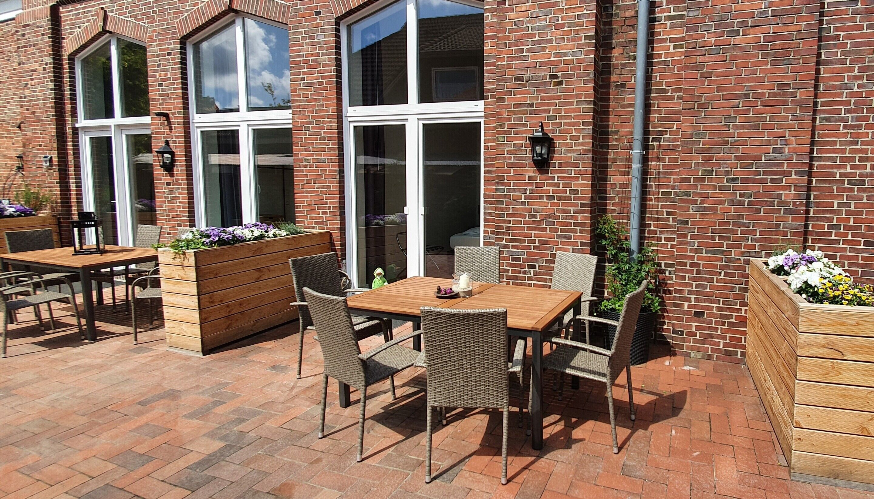 Eine Terrasse im Freien mit Ziegelmauern, Holztischen, Korbstühlen und Blumenkübeln.