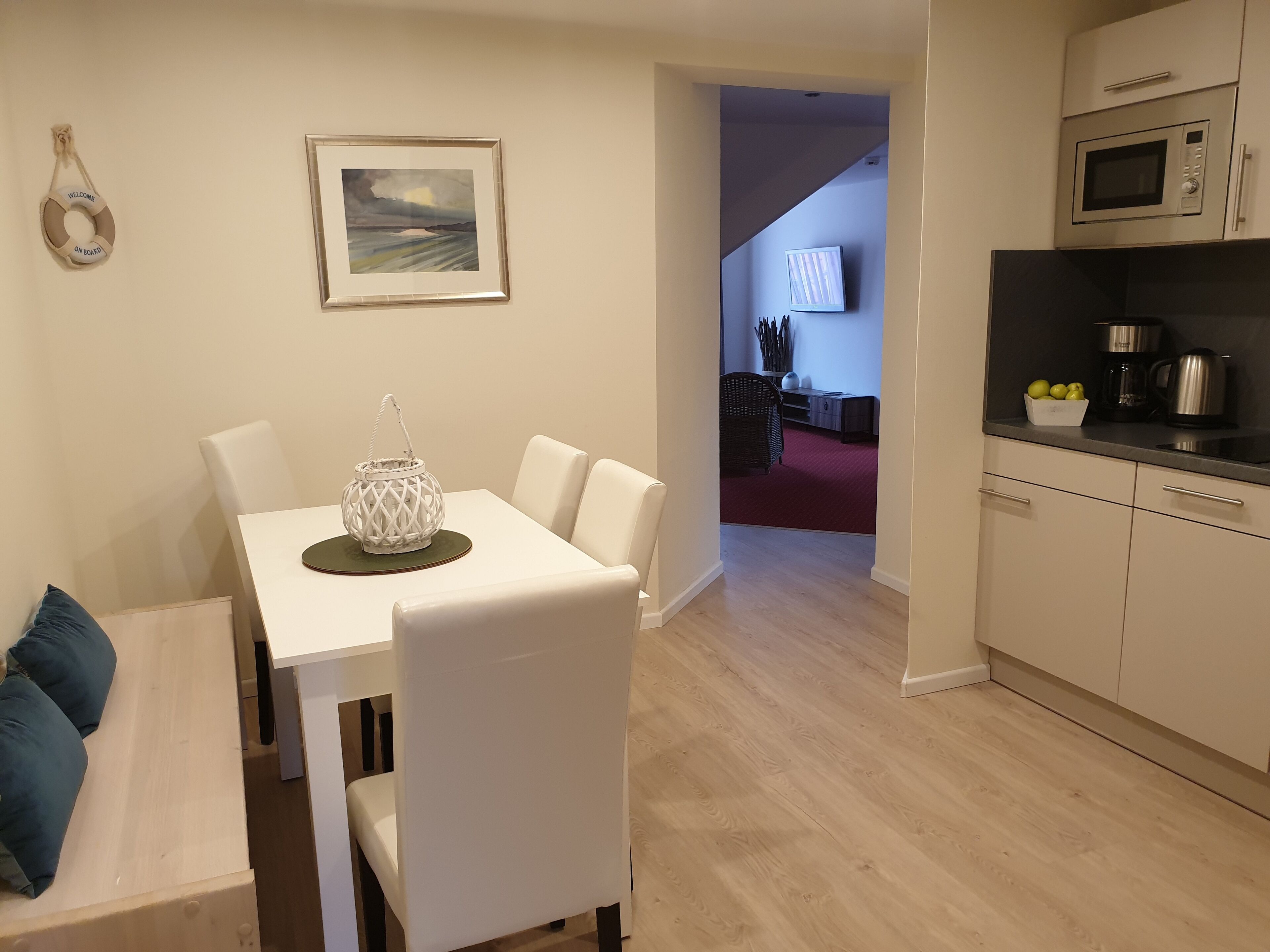 Cocina moderna con armarios blancos, microondas y cafetera. Hay un comedor con mesa y sillas blancas, y un cuadro en la pared.