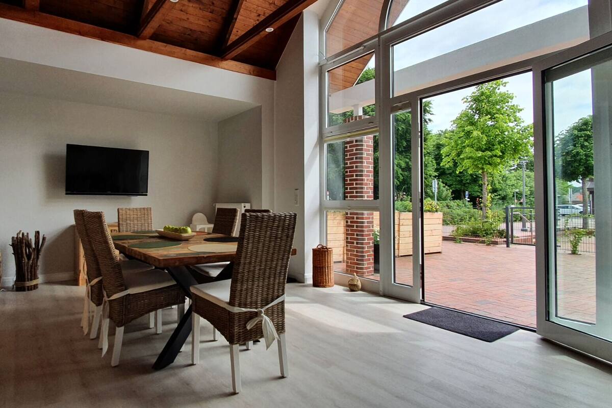 A dining area with a large table, wicker chairs, a television on the wall, and large windows offering a view of the outdoors.