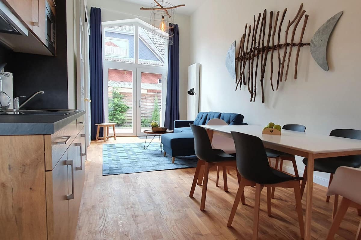 A modern kitchen with a built-in oven, a dining area with a white table and black chairs, and a large window with curtains.