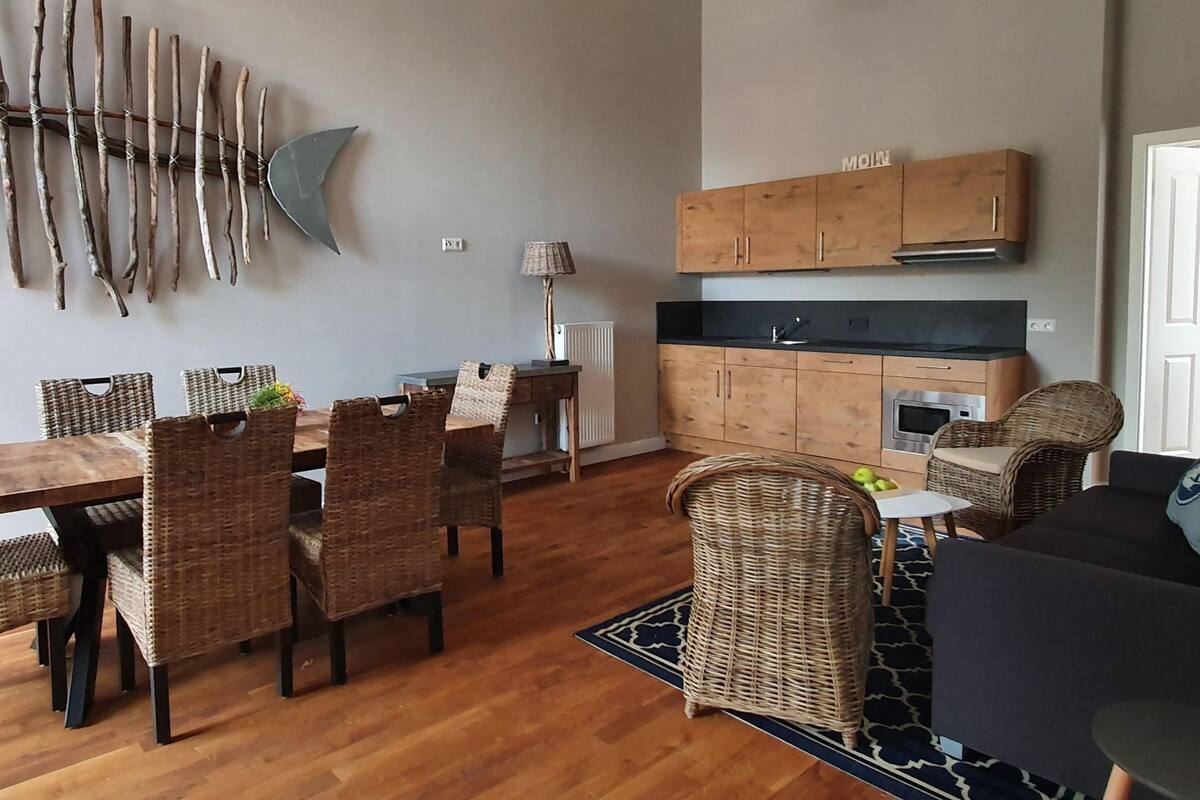 A modern kitchen with wooden cabinets and a black countertop. There is a dining area with wicker chairs and a wooden table.