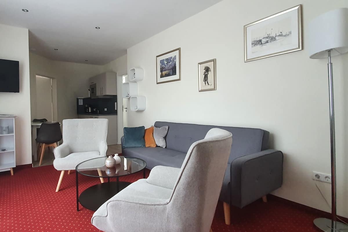 A living room with a grey sofa, a white armchair, a glass coffee table, and a TV mounted on the wall.