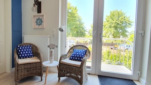 A room with two wicker chairs with anchor-patterned cushions, a small table, and a view of a balcony with a railing and parked cars. - Hotel Friesischer Hof (Hage)