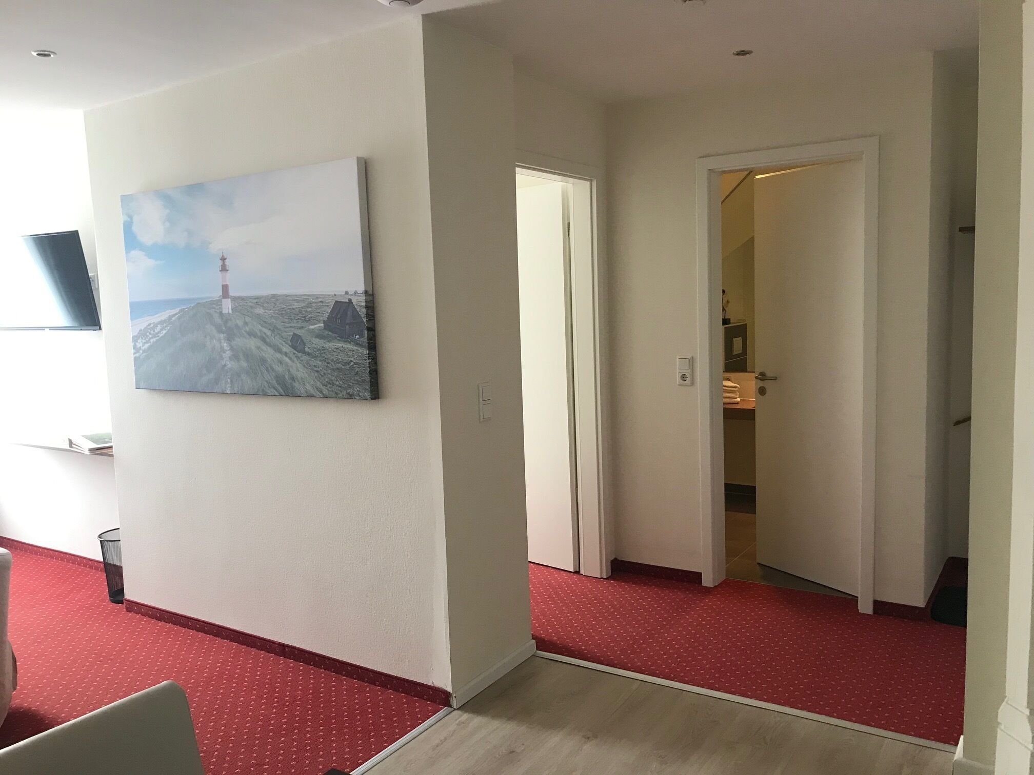 A hallway with a red carpet, a painting of a lighthouse, and a door leading to a room with a bed and a nightstand.