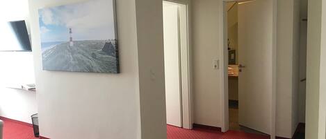 A hallway with a red carpet, a painting of a lighthouse, and a door leading to a room with a bed and a nightstand.