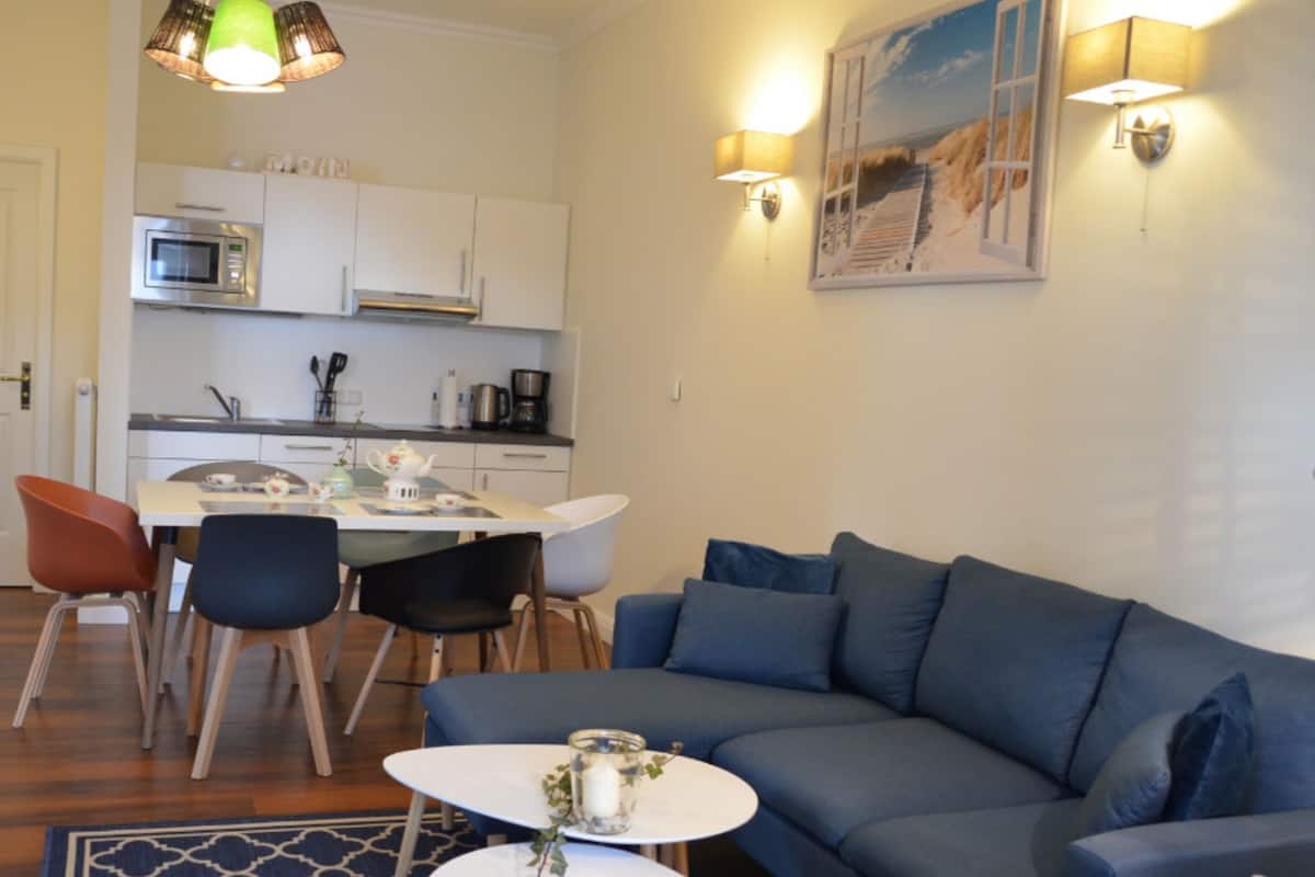 A modern living room with a blue sofa, a dining table with chairs, a kitchen area with white cabinets, and a painting on the wall.