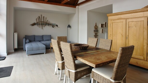 A dining area with a wooden table, wicker chairs, and a blue sofa in the background. - Hotel Friesischer Hof (Hage)