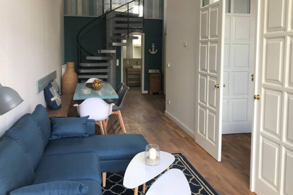 A compact living space with a blue sofa, a dining table with chairs, a spiral staircase, and a white door.