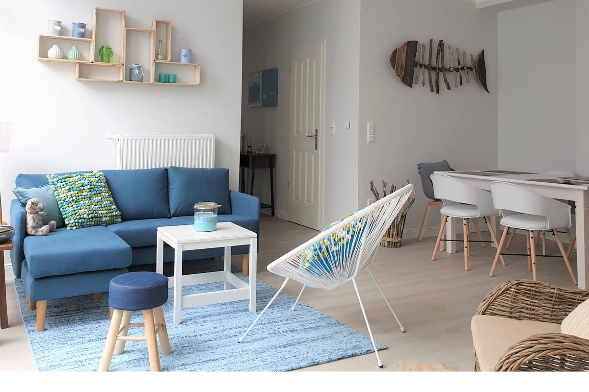 A modern living room with a blue sofa, a white dining table, and a white chair. There is a small white table with a blue container on it.