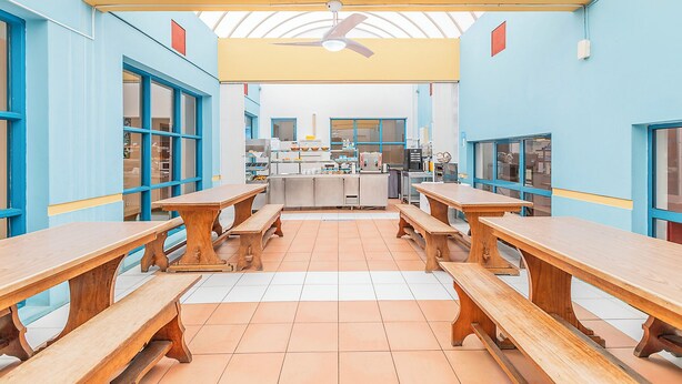 A cafeteria with wooden tables and benches, a ceiling fan, and a kitchen area in the background. - HI Faro- Pousada de Juventude - Hostel (Faro)