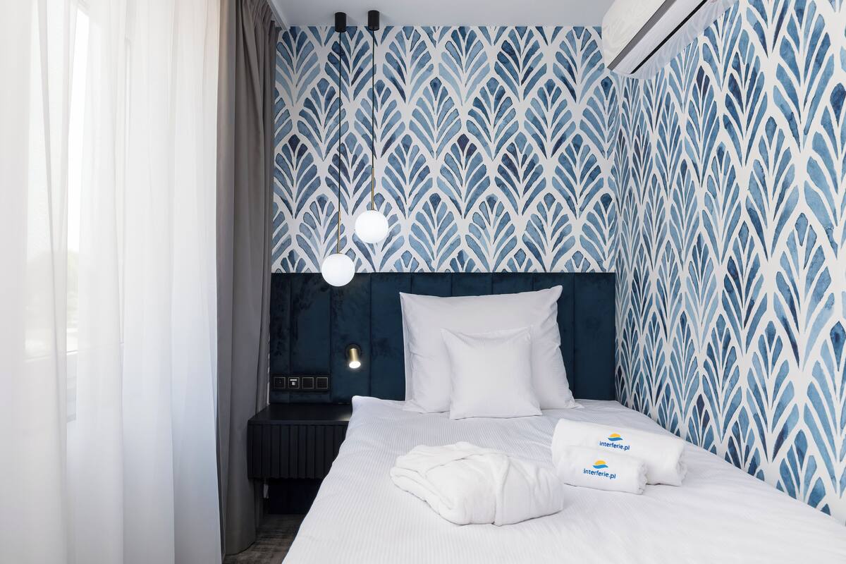A hotel room with a patterned wallpaper, a neatly made bed with white linens, a bedside table with a lamp, and a wall-mounted air conditioner.