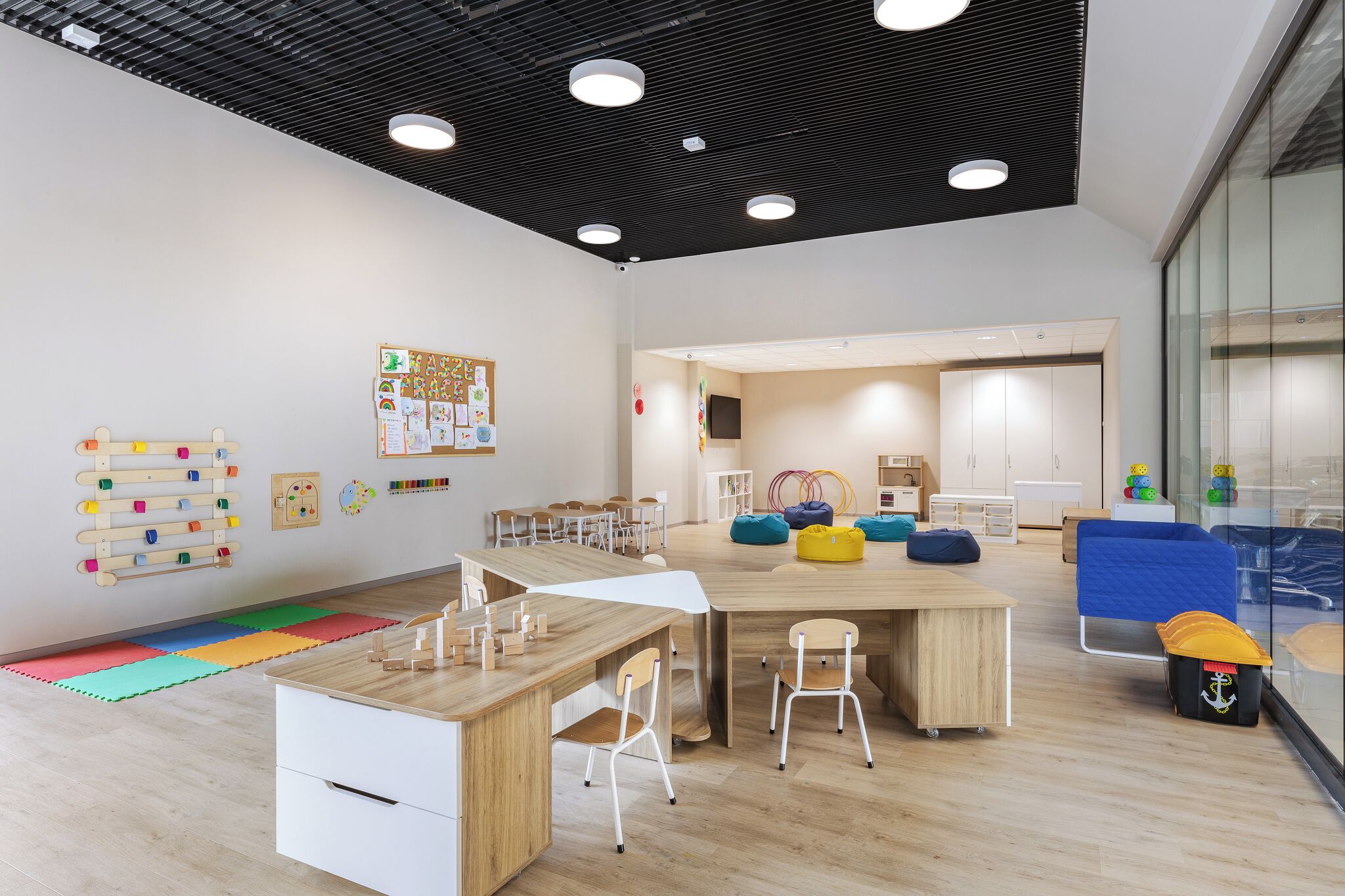 A modern classroom with a wooden desk, chairs, and colorful play mats on the floor.