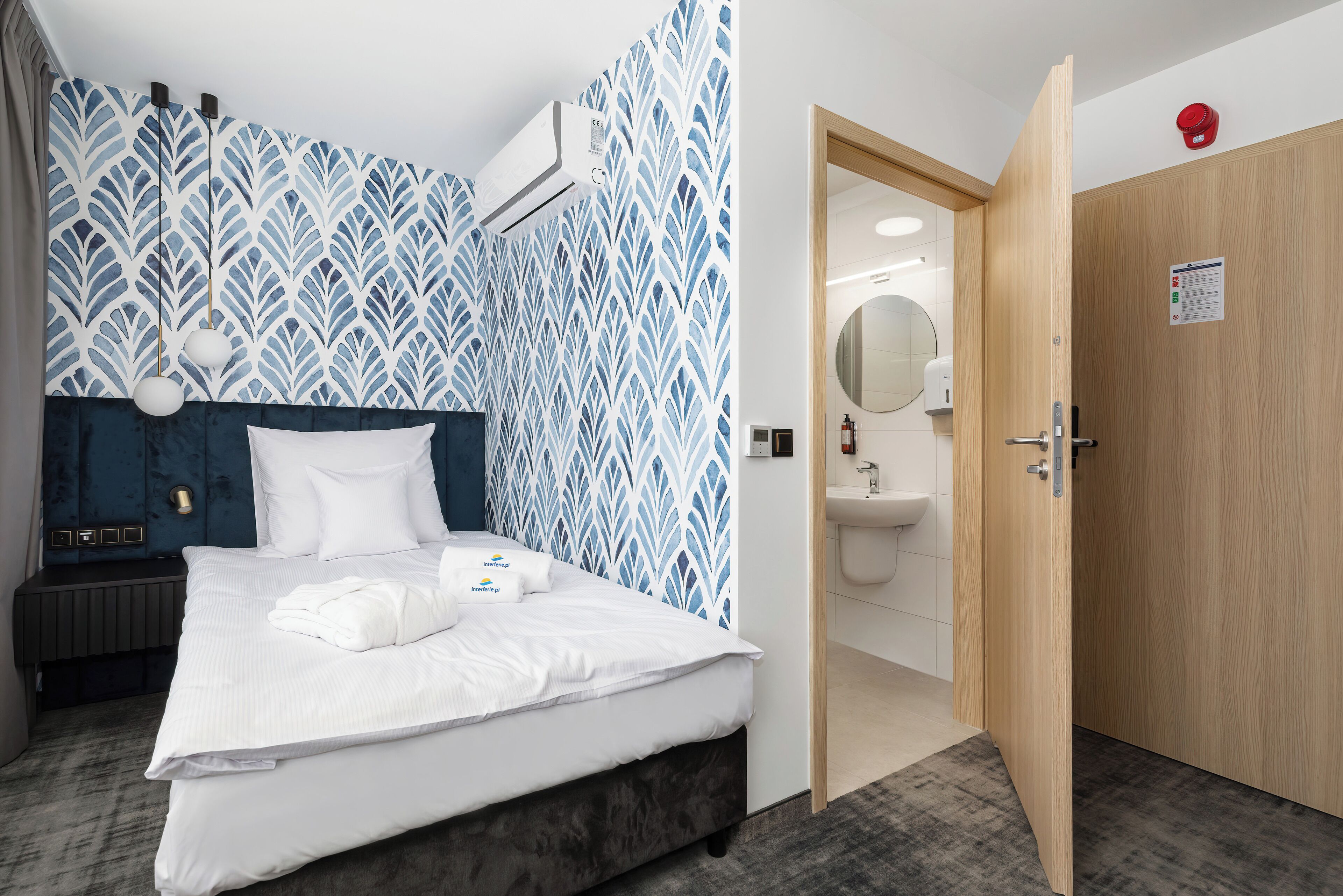 A modern hotel room with a large bed, a blue and white patterned wall, a wooden door, and a visible bathroom.