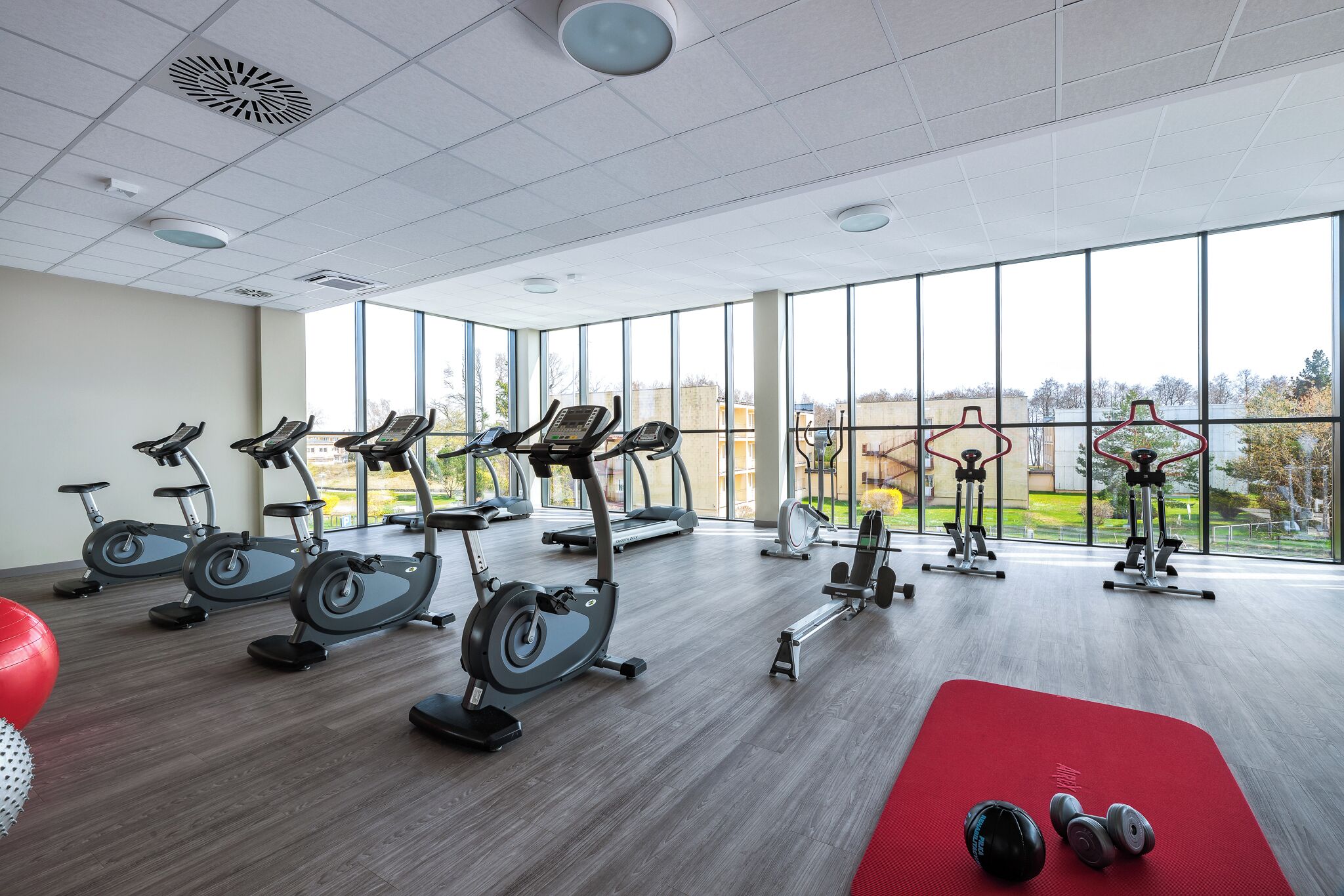 Une salle de sport équipée de nombreux appareils d’exercice, d’un tapis rouge et de grandes fenêtres donnant sur l’extérieur.