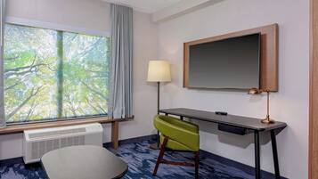 A hotel room with a large window, a desk, a chair, a lamp, and a flat-screen TV.