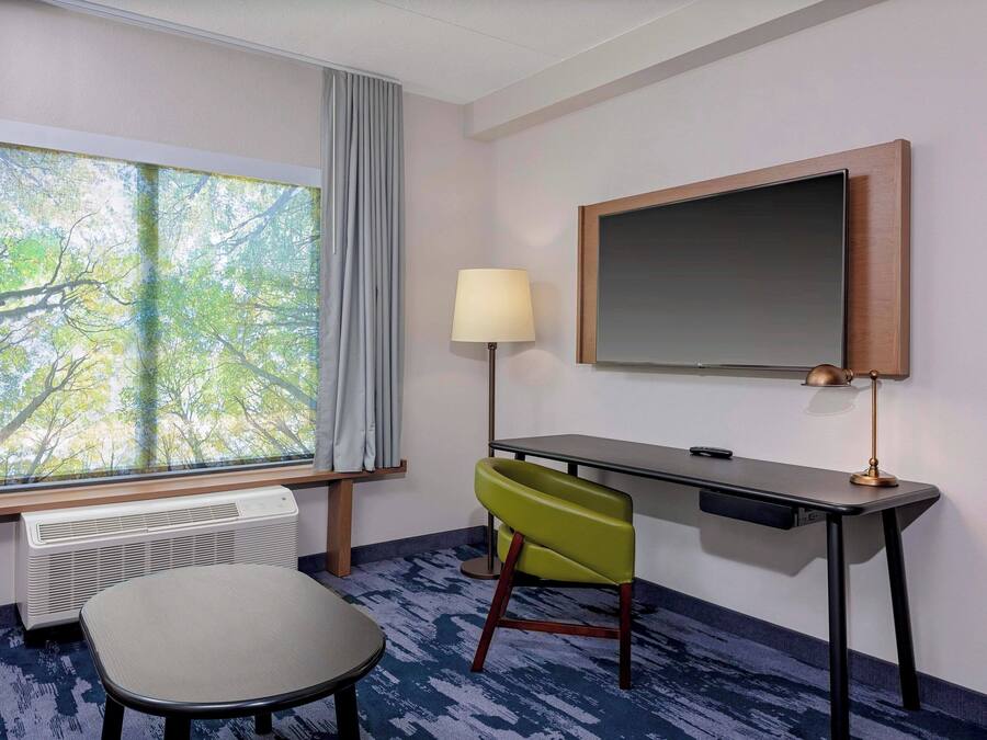 A hotel room with a large window, a desk, a chair, a lamp, and a flat-screen TV.