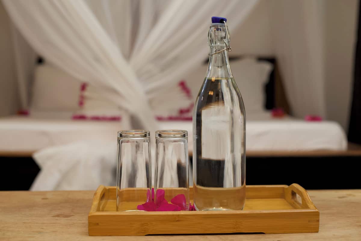 Una bandeja de madera con una botella transparente y dos vasos, una flor rosa, y una cama con ropa blanca de cama al fondo.