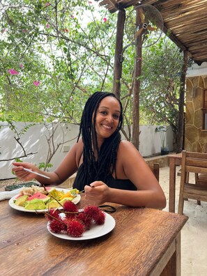 A person sitting at a wooden table with a plate of fruit and a fan in a covered outdoor area with trees and a stone wall.