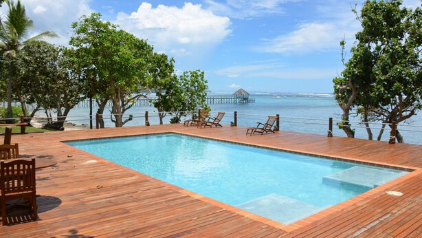 A rectangular pool with wooden decking, surrounded by trees and overlooking a body of water with a pier in the distance. - Fijian Oasis (Korolevu)