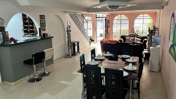 A spacious dining area with a central table set for four, surrounded by chairs. A bar area with a counter and stools is visible to the left. The room features a staircase leading to an upper level, large windows with a view, and a water dispenser on the right. - Tita´s House Bogotá (Bogotá)