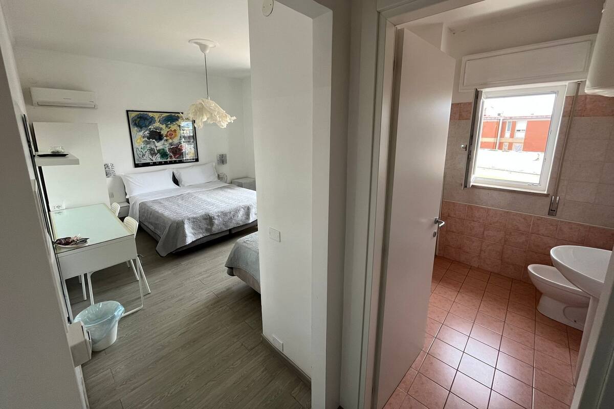 A hotel room with a bed, a desk, a chair, a trash bin, a painting on the wall, and a bathroom with a sink and toilet.