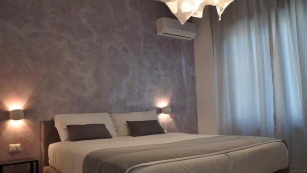 A bedroom with a large bed, two wall-mounted lights, an air conditioning unit, and a window with curtains. - F Rooms (Naples)