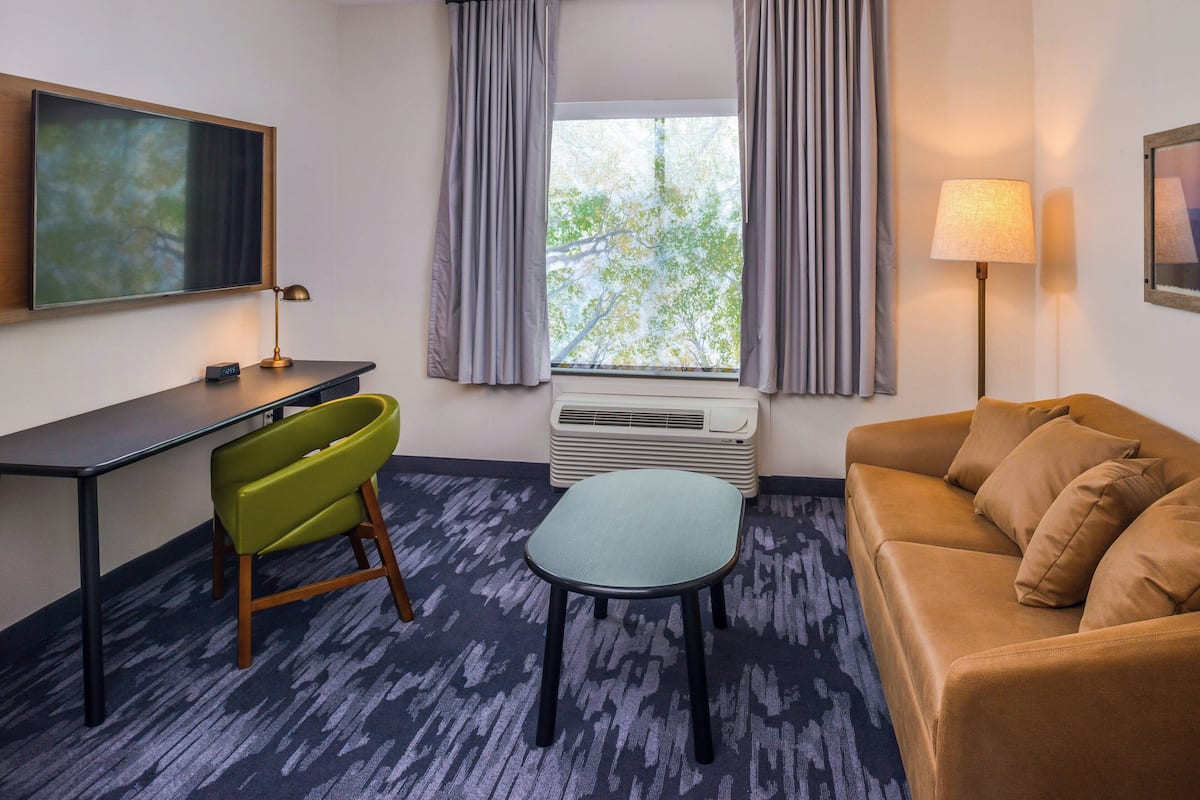 Habitación de hotel con escritorio, una silla verde, sofá marrón, una mesita, lámpara de pie y ventana con cortinas.