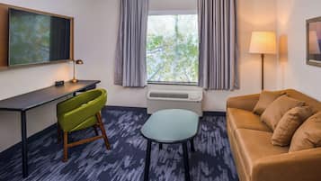 A hotel room with a desk, a green chair, a brown sofa, a small table, a standing lamp, and a window with curtains.