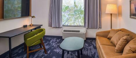 A hotel room with a desk, a green chair, a brown sofa, a small table, a standing lamp, and a window with curtains.