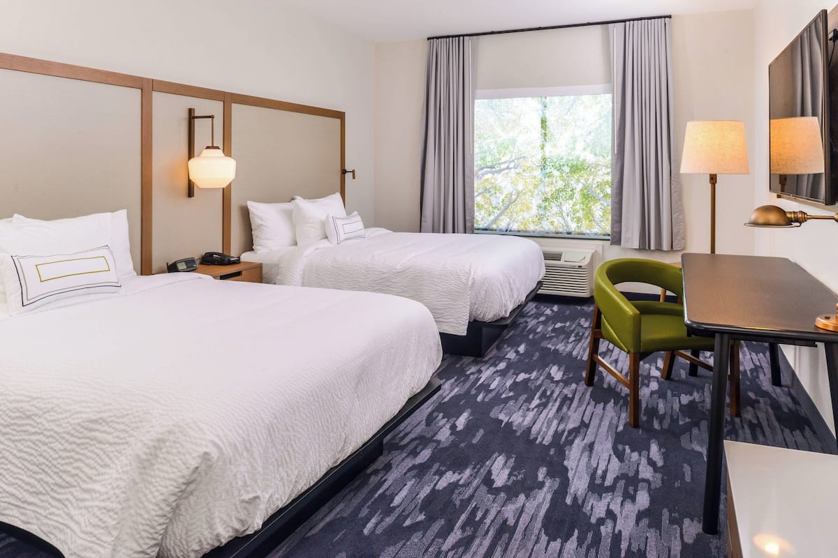 Habitación de hotel con dos camas, un escritorio, una silla y un gran ventanal con cortinas.