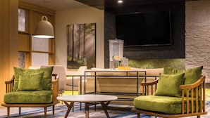Un área de descanso moderna con dos sillones verdes, una mesa redonda de centro y un sofá. Un televisor de pantalla plana grande está montado en la pared, y hay un cuadro de un bosque.