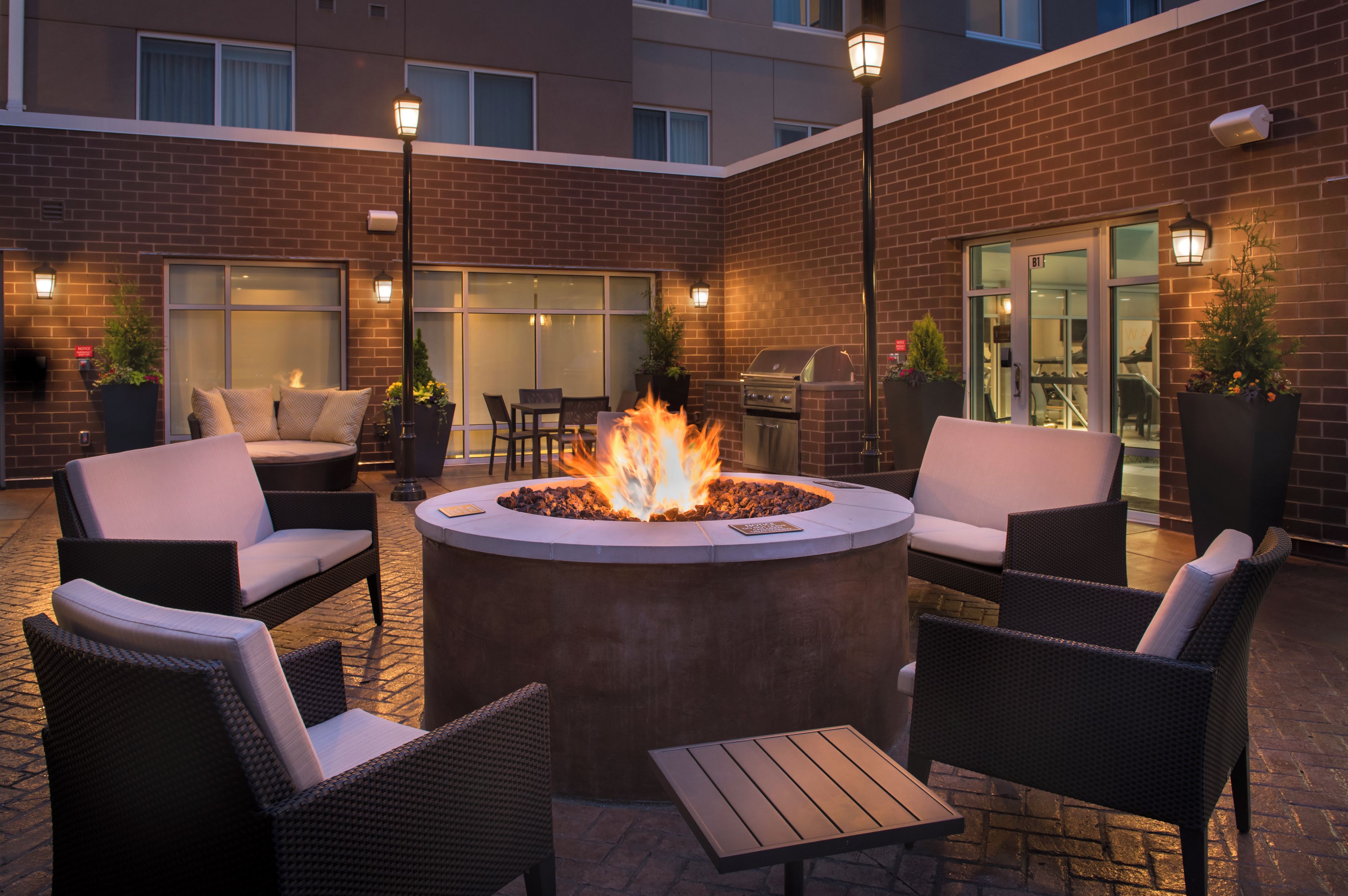 An outdoor seating area with a circular fire pit, wicker furniture, and a brick building in the background.