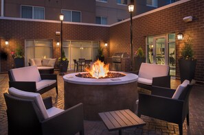 An outdoor seating area with a circular fire pit, wicker furniture, and a brick building in the background.
