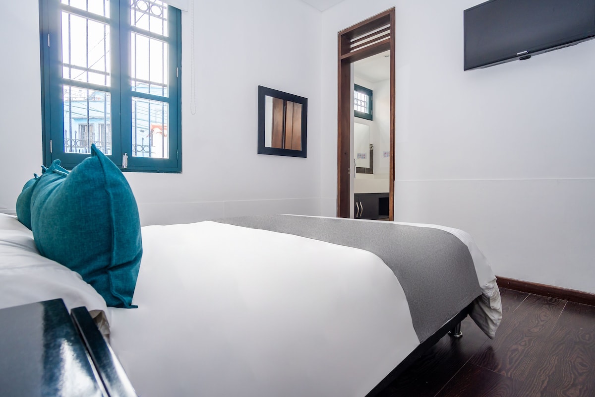 Una habitación de hotel con cama, ventana, espejo y televisor.