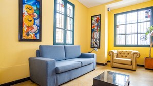 A living room with a blue sofa, a beige armchair, and a coffee table. There are two paintings on the walls and a window with a view of the outside. - Ayenda La Tua Casa (Bogotá)