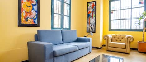 A living room with a blue sofa, a beige armchair, and a coffee table. There are two paintings on the walls and a window with a view of the outside.