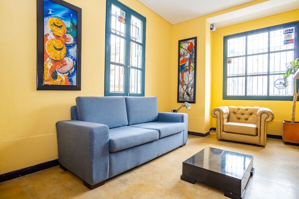 Una sala de estar con un sofá azul, un sillón color crema y una mesa de centro. Hay dos cuadros en las paredes y una ventana con vistas al exterior.