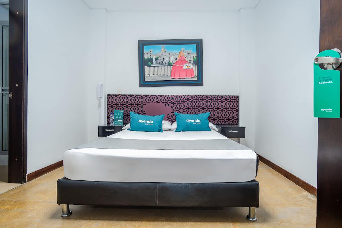 A hotel room with a large bed, two pillows with the hotel's logo, a framed picture on the wall, and a sign on the door.