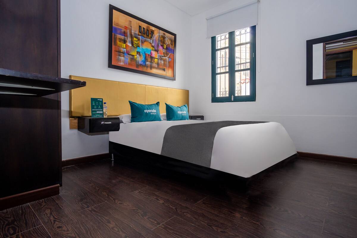 A hotel room with a large bed, a headboard, a nightstand, a desk, a window, and a painting on the wall.