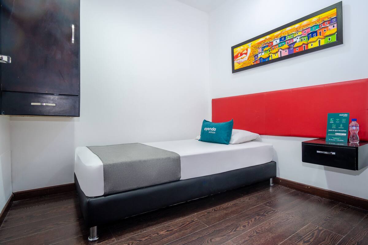 A hotel room with a bed, a red headboard, a wall-mounted TV, a shelf with toiletries, and a colorful painting.