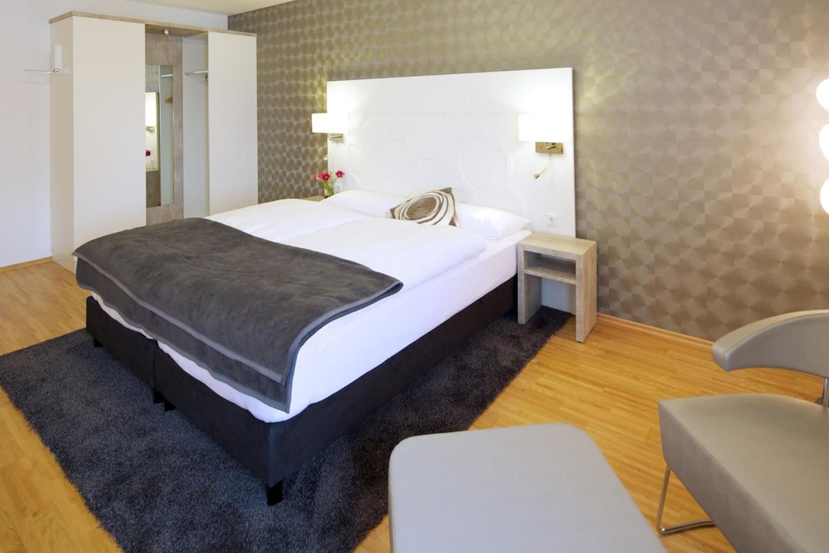 A modern hotel room with a large bed, bedside tables, a chair, and a view of the hallway.