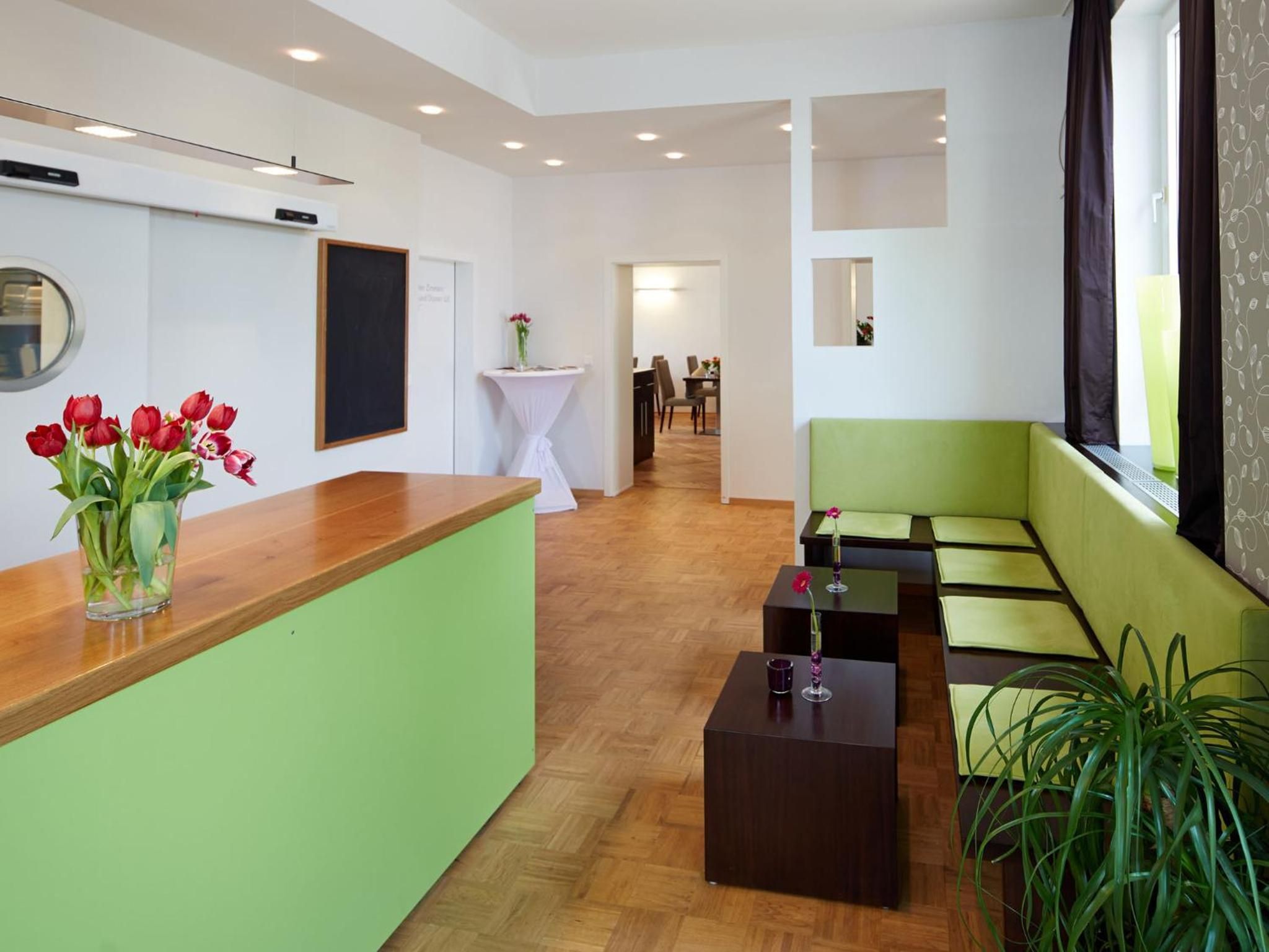 Eine Hotellobby mit einer Rezeption, einem grünen Sofa, einem kleinen Tisch mit einer Blumenvase und einer Topfpflanze.
