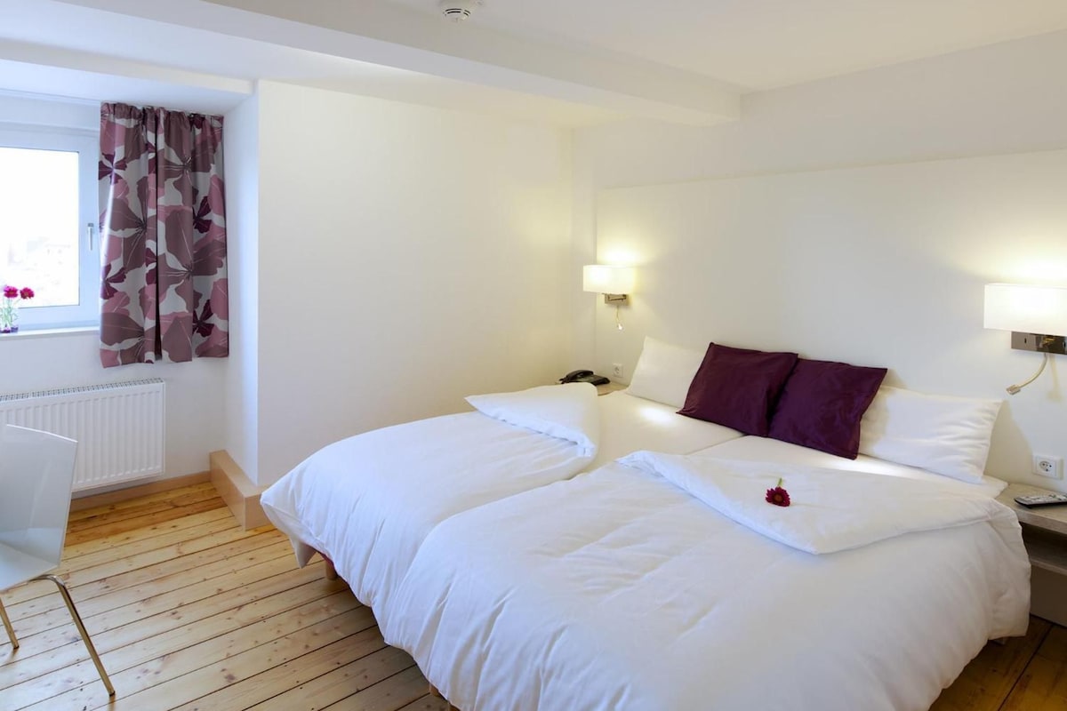 A hotel room with two beds, a desk, a radiator, and a window with curtains.