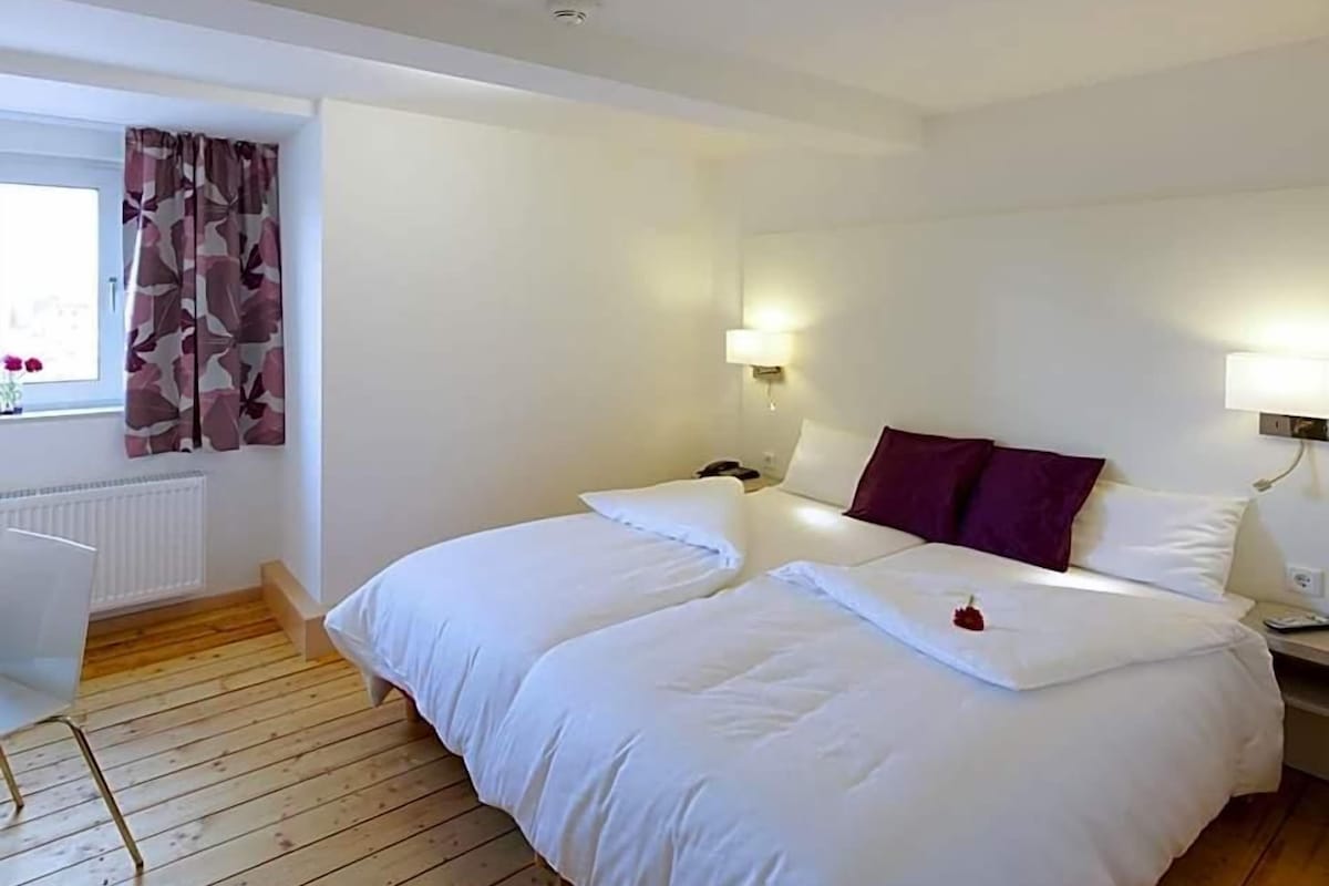 Una habitación de hotel con dos camas, una ventana con cortinas, un radiador y lámparas fijadas a la pared.