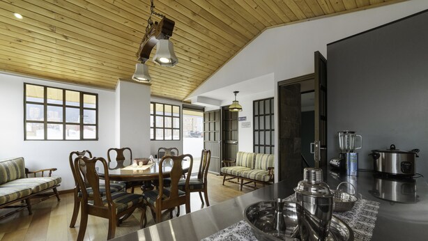 A spacious kitchen with wooden ceiling, a dining area with wooden chairs and a sofa, and a view of the city through large windows.