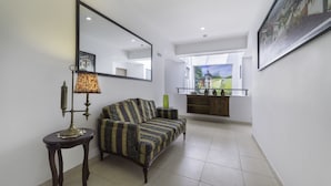 A living room with a sofa, a table with a lamp, a mirror, and a kitchen area in the background.