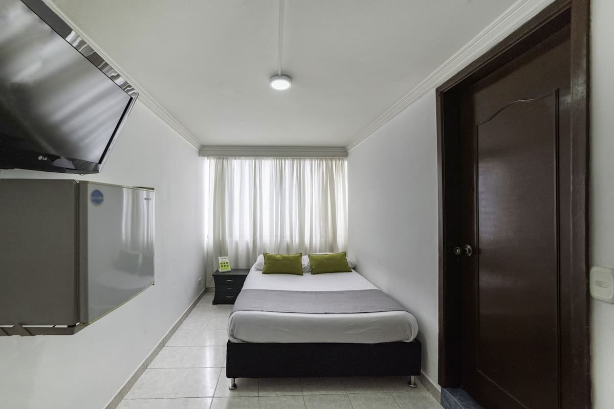 A hotel room with a bed, a TV mounted on the wall, a desk with a lamp, and a door.