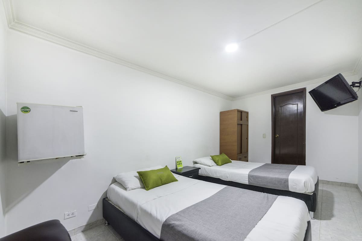 A hotel room with two single beds, a TV mounted on the wall, a wooden wardrobe, and a white air conditioner.